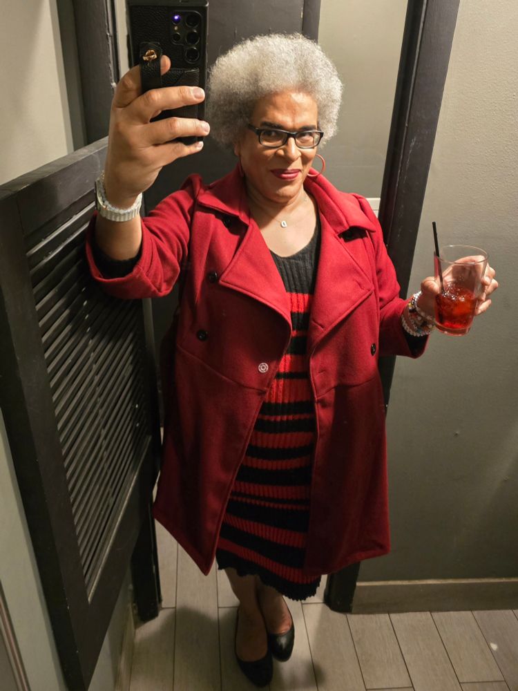Dana taking a restroom mirror selfie, holding a vodka cranberry cocktail, wearing a red and black striped sweater dress underneath a double breasted red overcoat.