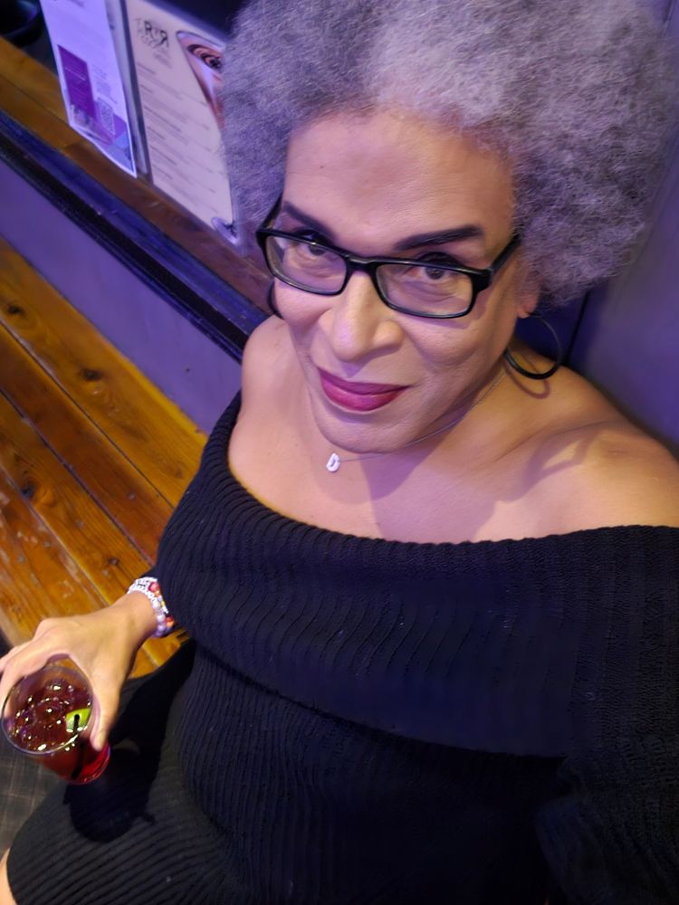 Selfie of Dana in a black cold shoulder sweater dress, holding a vodka cranberry, sitting on a bar counter.