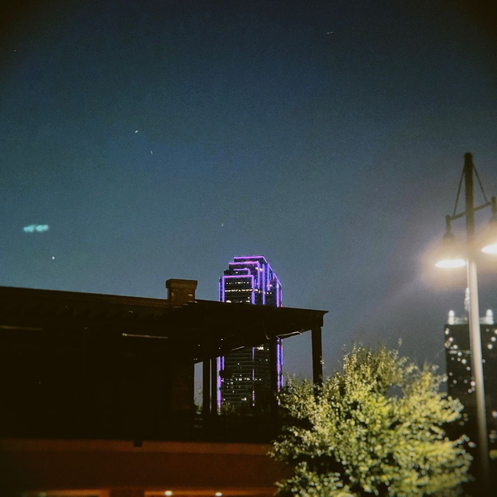 dallas skyline at night, one building visible, lit up purple, tree and building in foreground 