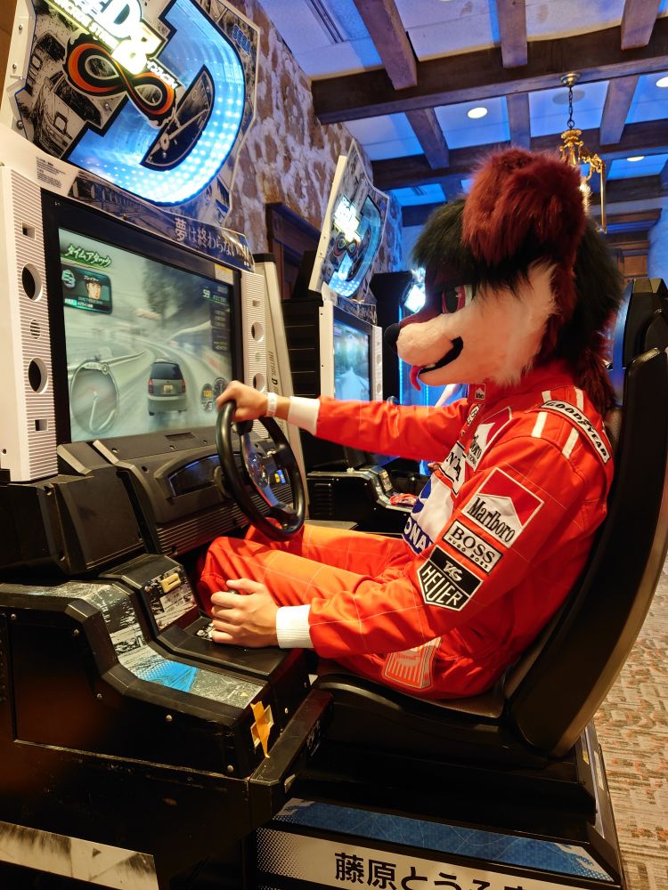 A dog fursuiter in a full racing suit playing Initial D 8 in the Stratosfur game room