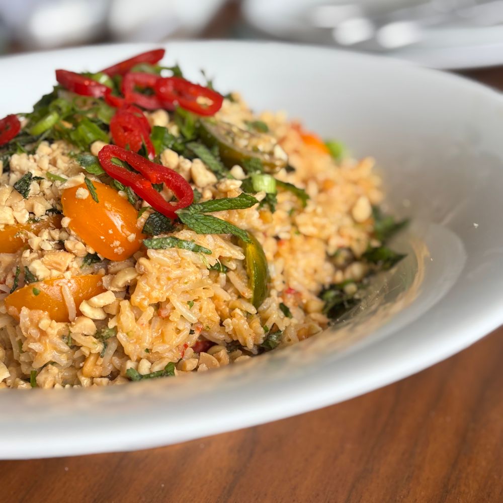 A close-up of a vibrant rice salad served in a white bowl. The dish includes fluffy rice mixed with chopped fresh herbs like mint and coriander, finely chopped peanuts, sliced red chillies, and halved rainbow cherry tomatoes. The salad is dressed with a spicy, tangy dressing, giving it a rich orange hue.
