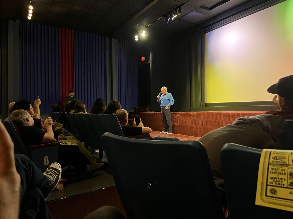 Peter Weller speaking on a microphone in front of the screen at the New Beverly.