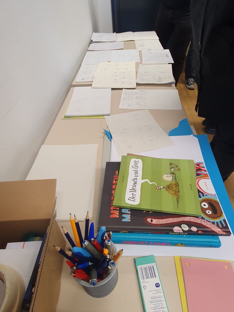 Zu sehen ist ein Tisch, auf dem erste Comicentwürfe und Zeichenübungen liegen, zudem einigen Utensilien und auch Beispielcomics.