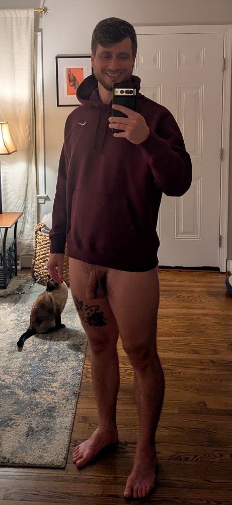 James taking a selfie wearing a burgundy hoodie and smiling. The frame shows his entire person from head-to-toe, which reveals he is naked other than wearing the hoodie. His cat, Canela, sits on a carpet in the background.