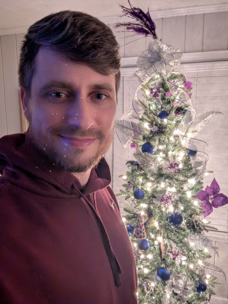 James taking a selfie wearing a burgundy hoodie in front of the holiday tree and smiling. The frame is from his chest up.