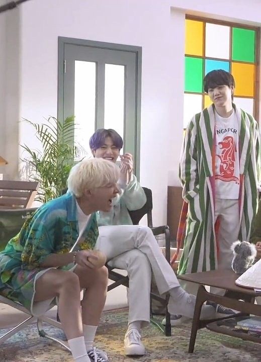 J-Hope is sitting in a chair laughing hard with Jungkook right next to him. Suga is standing and looking at them fondly.