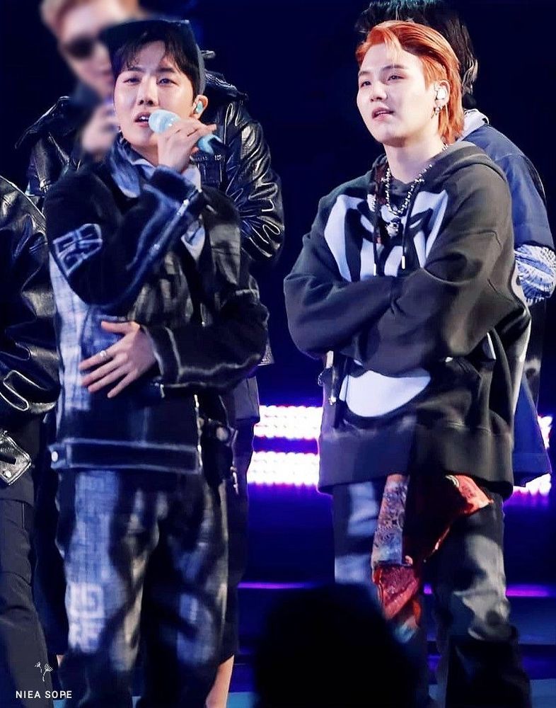 J-Hope and Suga are both standing on stage with their arms crossed