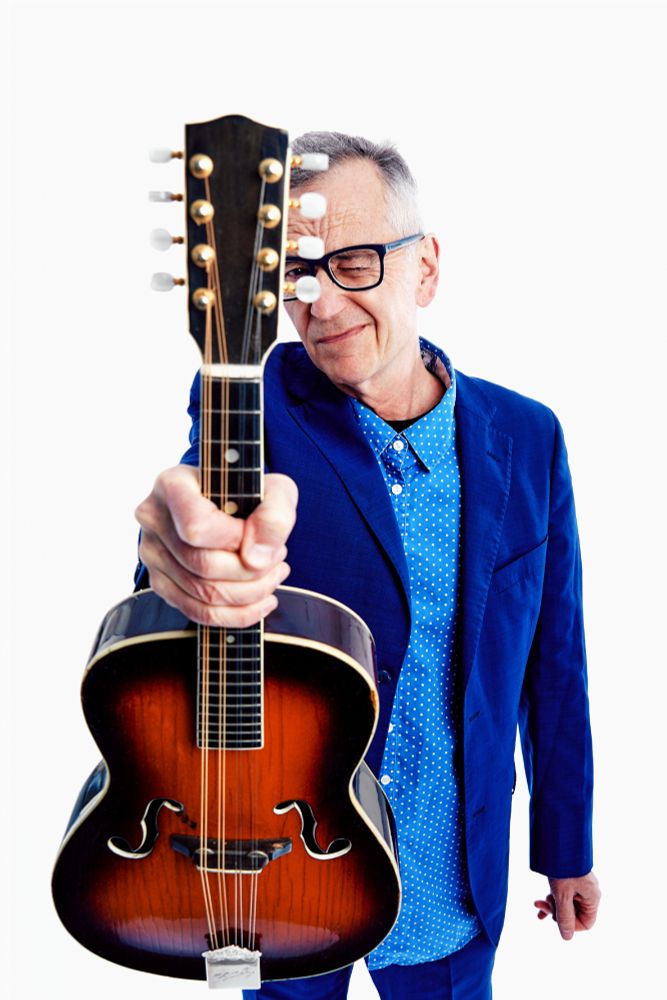 A man with short grey hair and glasses, wearing a blue jacket and a blue shirt with white spots, has his right arm outstretched towards the camera and is holding a mandolin. That man is John Hegley.