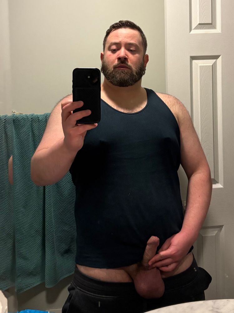 A mirror selfie from mid-thigh and up of me wearing a black tank top, and partially dropped sweatpants; my balls are flopped over the waistband and I’m tuggin my hard lil uncut dick