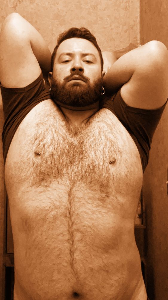 A sepia tone selfie of me (a bearded and hirsute chubby gay man) pulling my shirt back behind my head to expose my torso. 