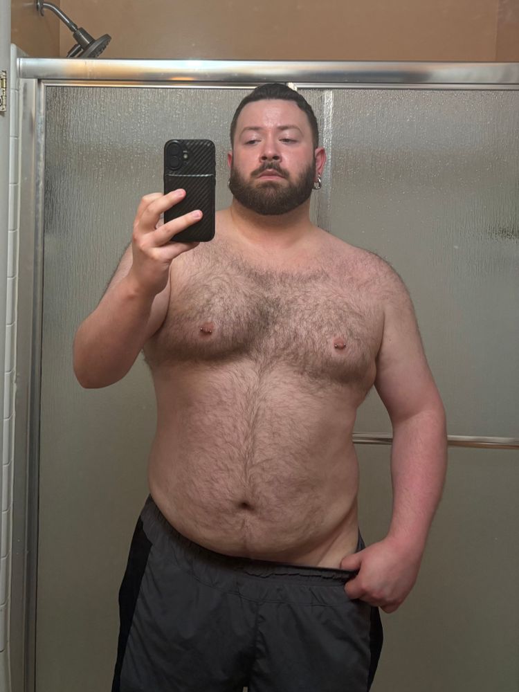 A shirtless mirror selfie showcasing a sleepy me and my hairy chubby torso. I’m wearing gray and black gym shorts and I’m standing in a before a cheap sliding shower door in a slightly dingy bathroom. 