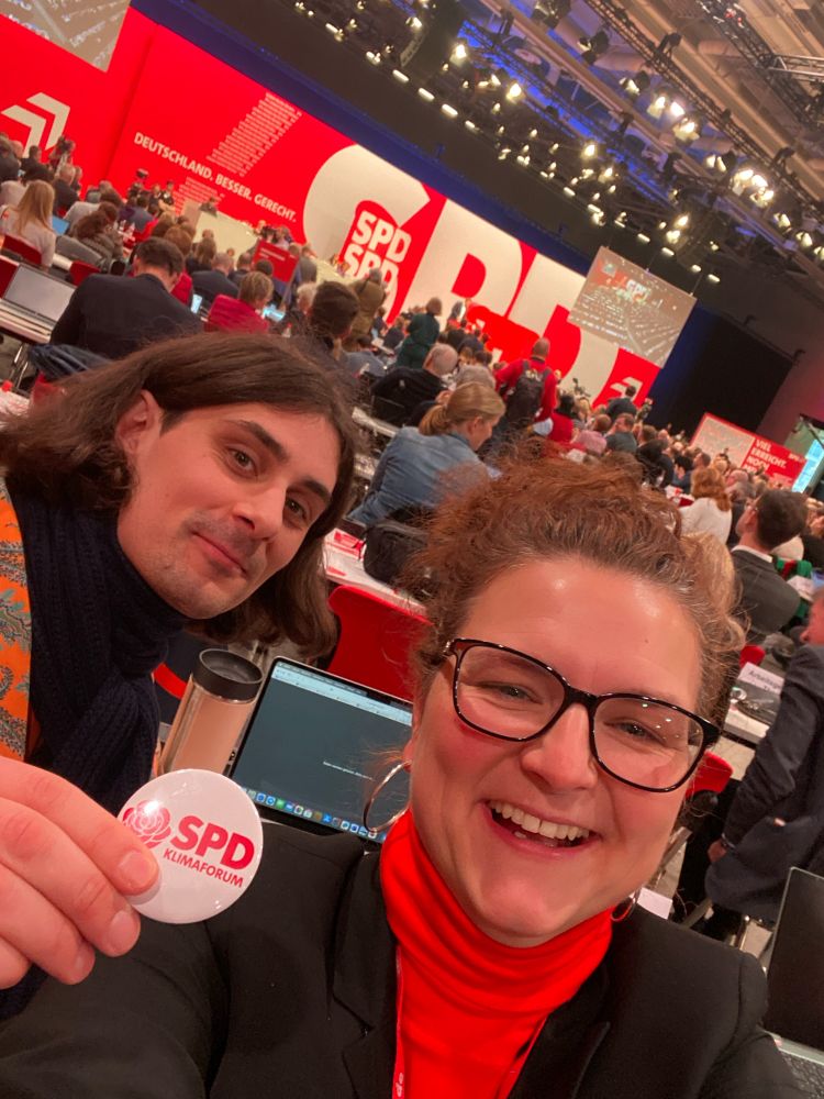 Ein Mann und eine Frau sitzen in einer großen Halle auf dem Bundesparteitag der SPD und halten einen Button mit der Aufschrift SPD-Klimaforum ins Bild.