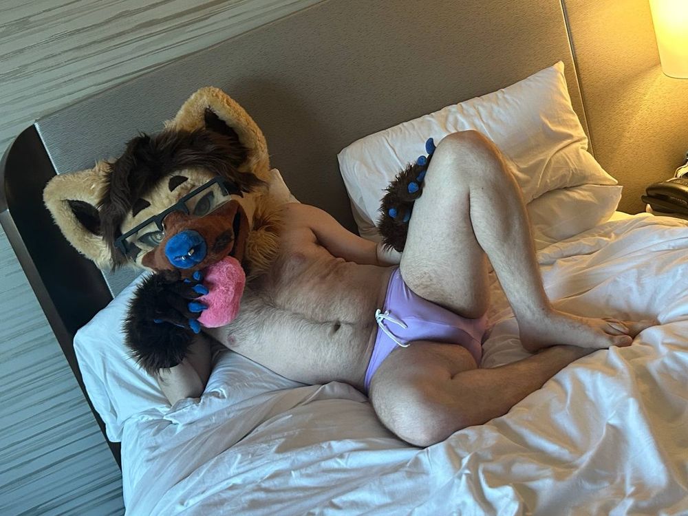 Suggestive picture of the hyena partial fursuiter laying in a bed seductively posing with a prop pastry in mouth