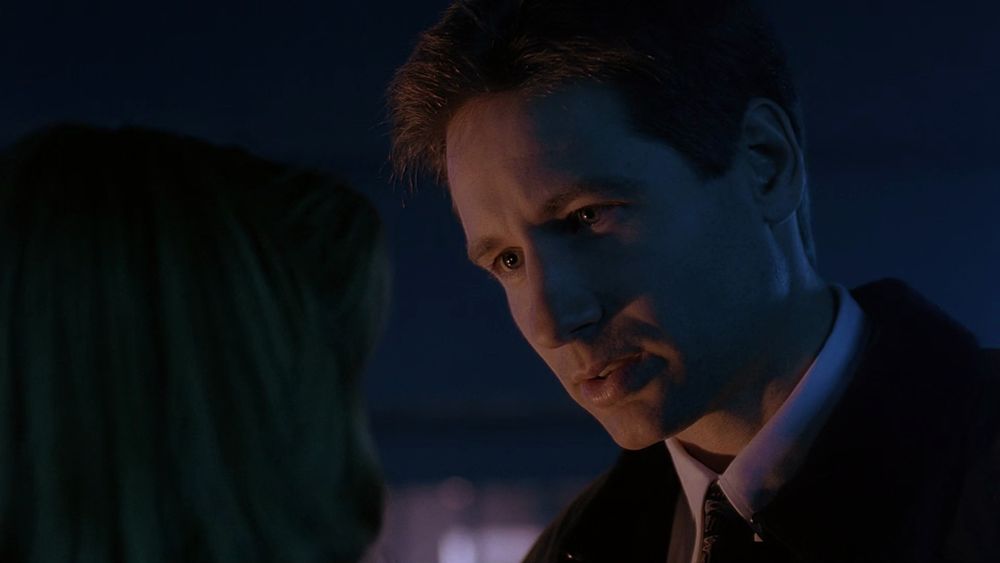 David Duchovny, a white brunette man, as Fox Mulder. He is looking to the bottom left of the screen at Scully. 