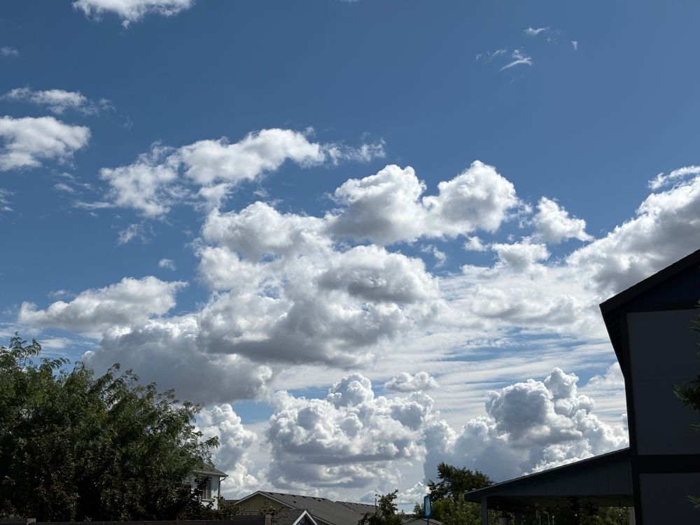 A photo of clouds against a blue sky. Trees and rooftops can be seen toward the bottom of the image.