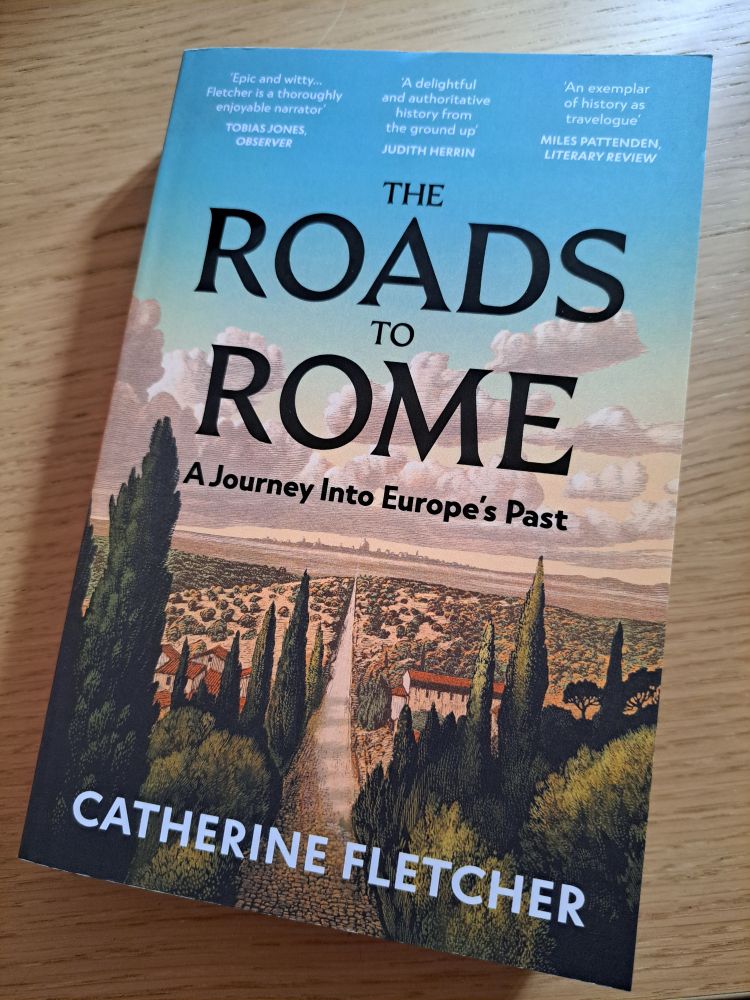 Paperback book with cover showing a long straight road between pine trees leading to a distant city beneath blue sky. Title of book is The Roads To Rome: A Journey into Europe's Past by Catherine Fletcher. Quotes on cover describe it as 'epic and witty - Fletcher is a thoroughly enjoyable narrator' (Tobias Jones, Observer), 'A delightful and authoritative history from the ground up' (Judith Herrin) and 'an exemplar of history as travelogue' (Miles Pattenden, Literary Review).