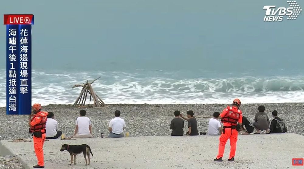 A screen capture from TVBS News showing the sea and a rocky beach with two rescue workers and a dog in the foreground and around 8 people watching for the tsunami to arrive.
