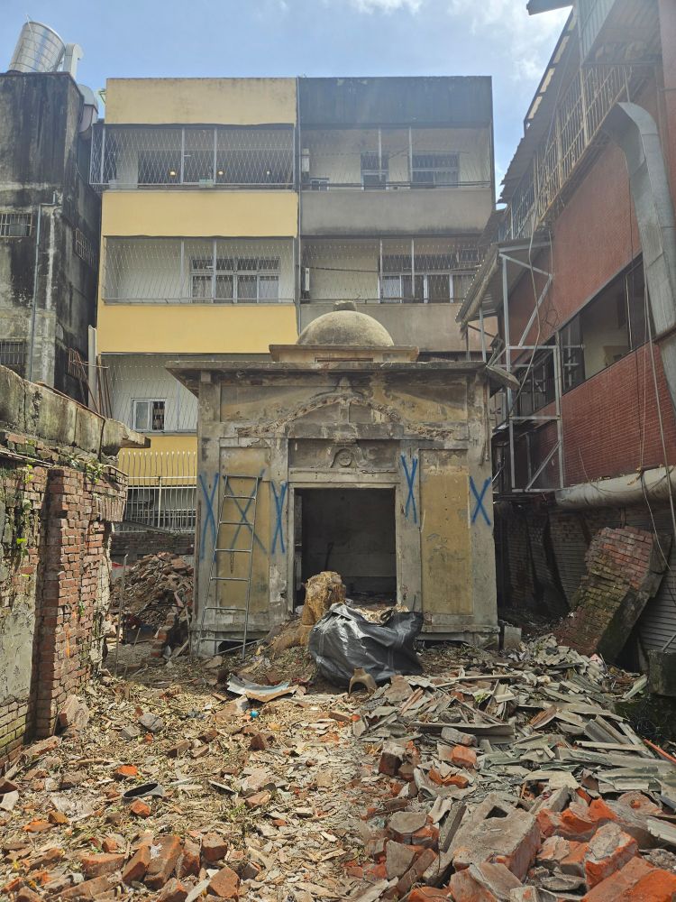 A mysterious reinforced concrete building recently unearthed in the back alleys of Taichung. It is square and minimalistic with a Buddhist swastika carved over the entrance. Debris is all around in the foreground.