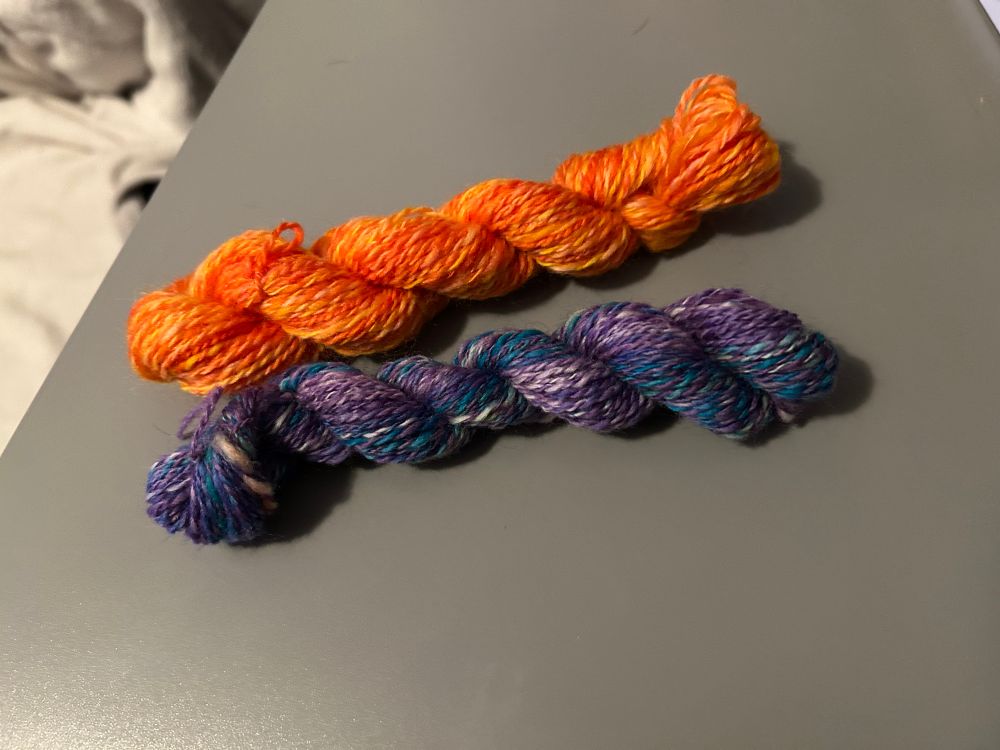 Two mini skeins of yarn. Top is a tonal orange in 39yd. Bottom is purple/blue variegated in 33yd. 
