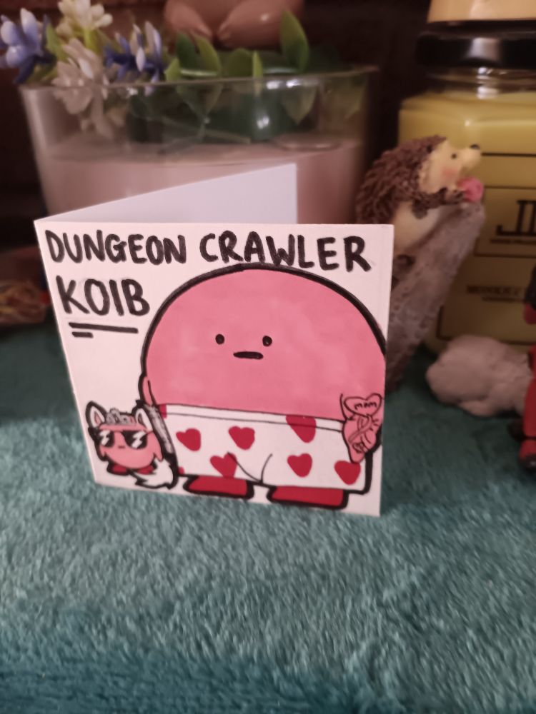 A small card with a represtation of the character Kirby dressed as the book protagonist Dungeon Crawler Carl titled Dungeon Crawler Koib. Beside him is a pink Kirbified cat drawing of Carl's partner Princess Donut.