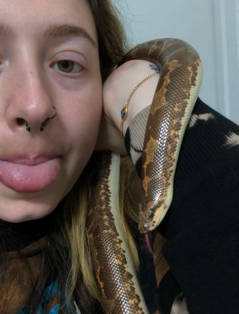 A close up photo of a fair skinned person holding the same snake up to their face - both are sticking their tongues out 