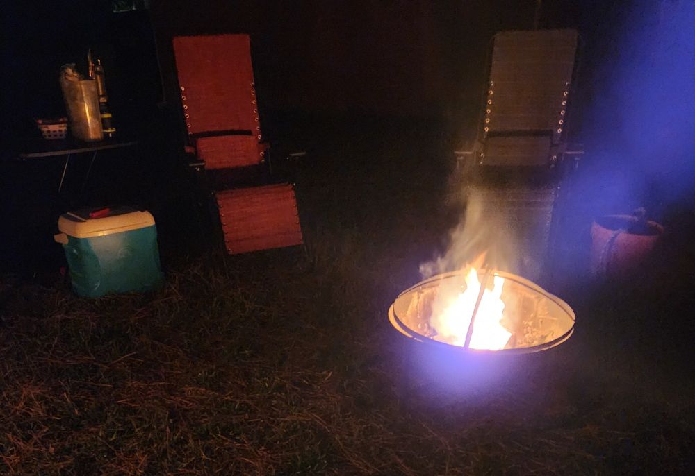 Back yard campfire near two chairs, a cooler of drinks (its actually water, for real), and smores supplies. For fire safety, near one of the chairs is a clay pot holding a water hose. Not shown and behind the cooler is a fire extinguisher. 