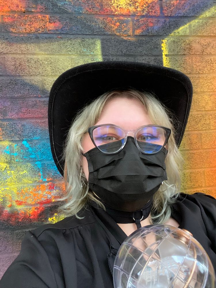 A selfie of me in front of the venue mural. My lightiny is visible in the lower right and I’m wearing my glasses and a black face mask. 