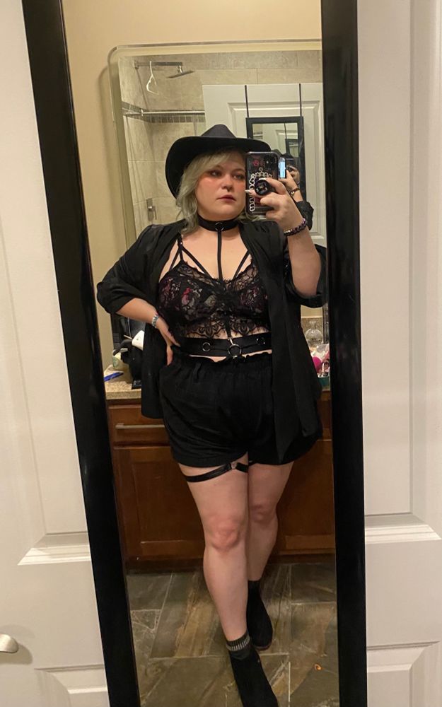 A mirror selfie taken of me in a full-length mirror in my bathroom to show off my concert outfit. I am wearing a lacy black crop-top over a rose-print bra; high waisted black shorts; a black body harness that goes from a collar around my neck to a waist belt, then to thigh garters that peek out under my shorts; an unbuttoned oversized black long sleeved blazer with cuffed sleeves; a black cowboy hat; and black ankle boots. I have shaggy silver shoulder-length hair, and exaggerated blushy makeup on. 
