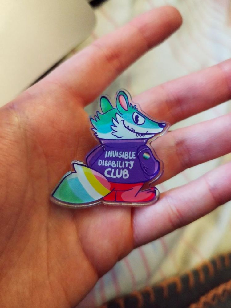 Eine Hand, auf der ein Anstecker liegt. Motiv auf dem Anstecker ist ein Cartoon-Waschbär mit Jacke, auf der "invisible disability club" steht. Der Waschbär ist in Farben der disability pride coloriert.