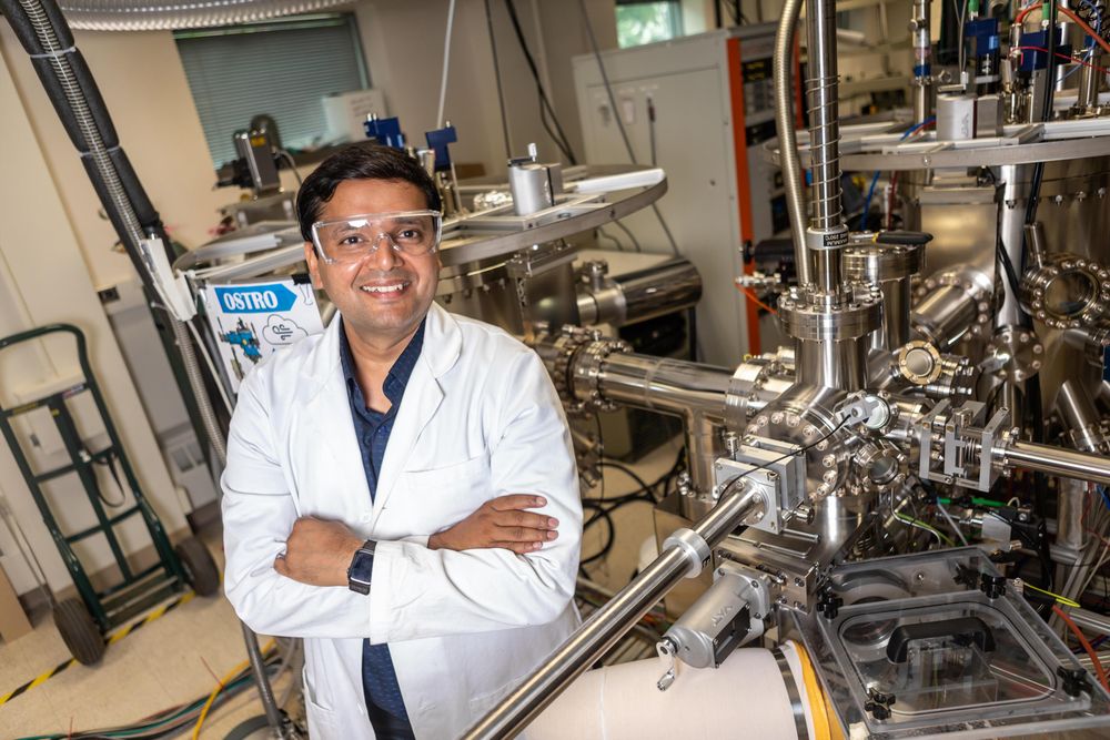 A person in a lab coat smiling and standing with arms crossed in a laboratory setting, surrounded by advanced scientific equipment.