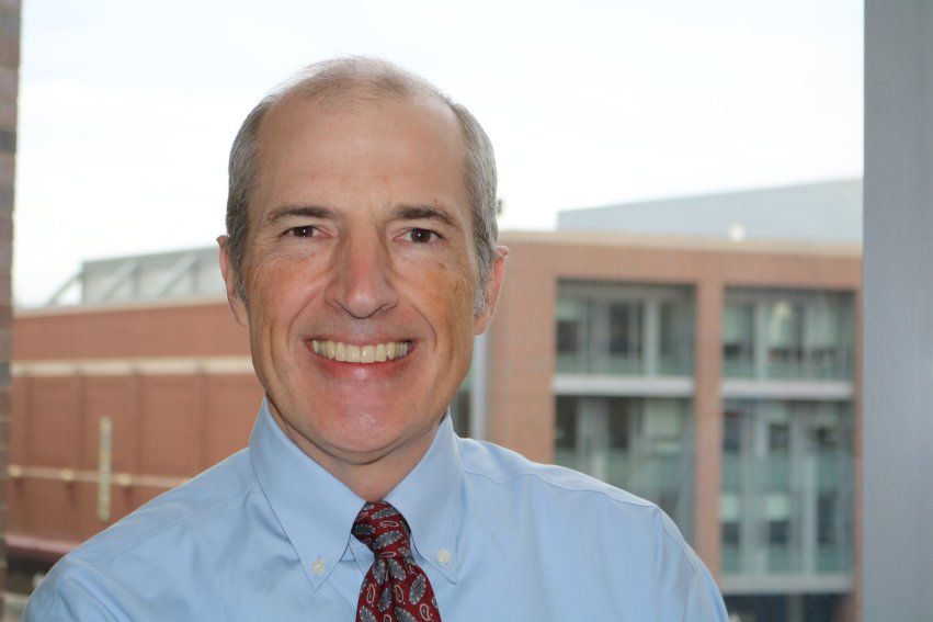 Portrait of an individual smiling, wearing a blue shirt and red tie, with an office building background.