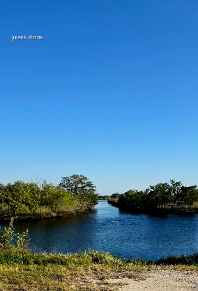 
A calm, narrow river stretches into the distance between low, bushy green trees and marsh grasses under a clear bright-blue sky. The shoreline in the foreground is sandy with patches of grass, and the water reflects the sky with gentle ripples. The text “julesk.store” appears in the upper left corner. 