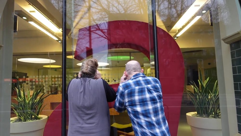 A man and a woman are peering through a large window of a building. Inside the window, there is a large "A" sculpture. On the left is Jenny, she is a white woman in a grey tunic, her hair is in an up-do. Kevin, a white man, in on the right, he is wearing a blue plaid shirt.