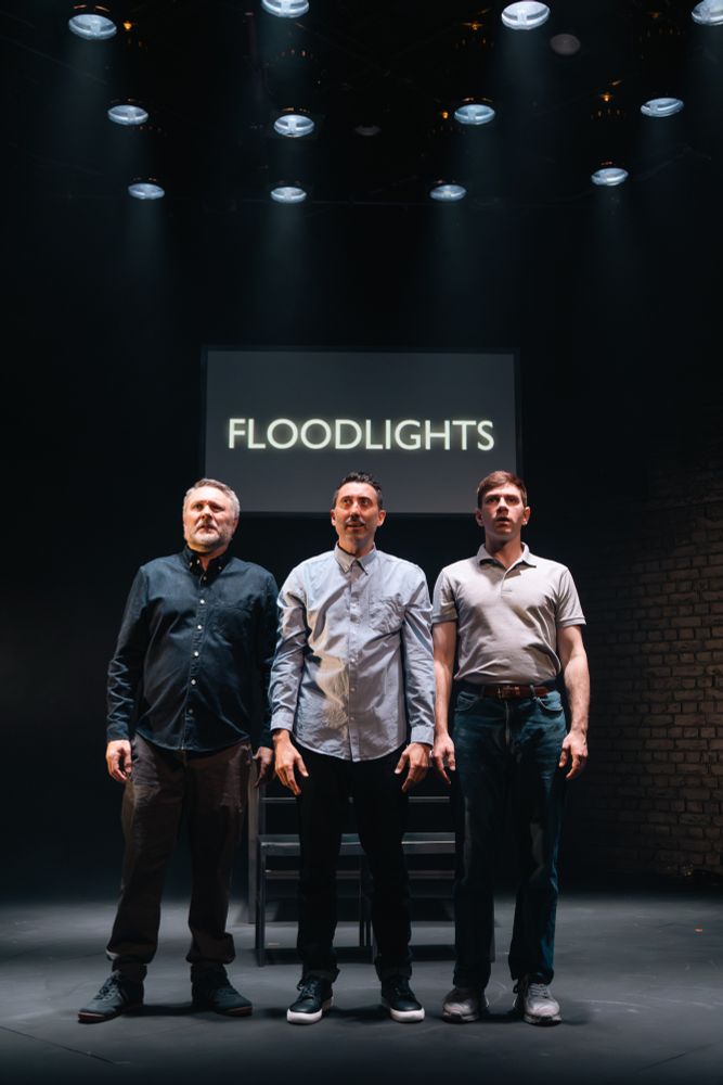 Three cast members stand shoulder to shoulder under flood lights. Caption screen behind reads in large font, Floodlights.