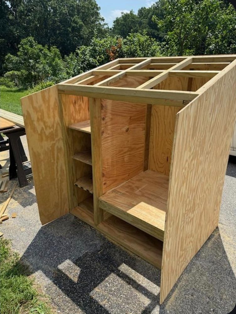 Wooden structure in the process of being built. It measures about 5 feet tall by 4 feet wide and 3 feet deep. It will hold a large mini fridge and has shelves for pantry food items.