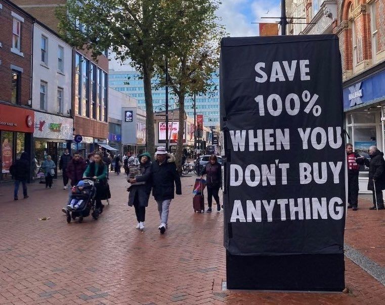 Photo of people walking in a high street.
A large black XR sign in the foreground reads "Save 100% when you don't buy anything*
