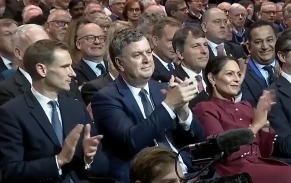 s/s from shared video showing the fixed smile on Priti Patel's face barely clinging on after she was excluded from the thanks. Two of the men around her are looking over at her as if to check her reaction.
