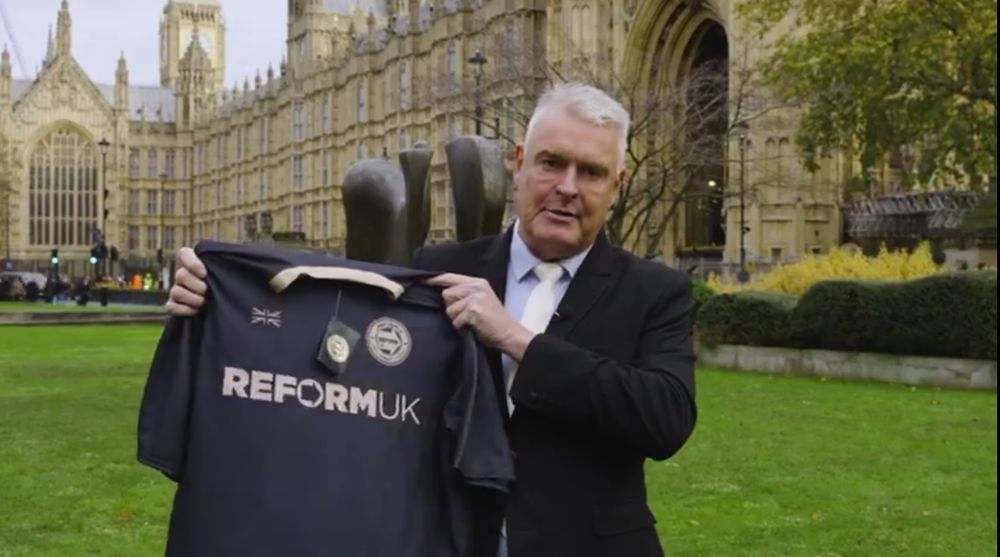 Tired and flabby Lee Anderson MP standing outside of the Houses of Parliament, holding up a Reform UK football black shirt with an uncomfortable expression