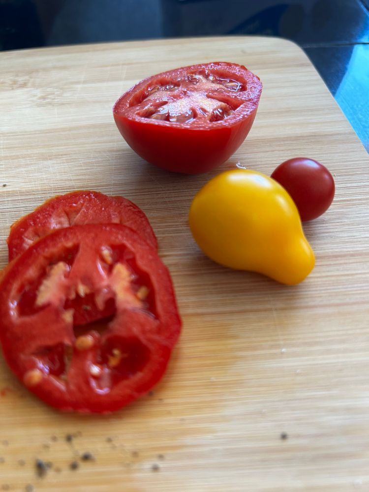Several tomatoes on a wooden cutting board - a medium sized red one that is sliced, a yellow cherub tomato, and a red cherub tomato.