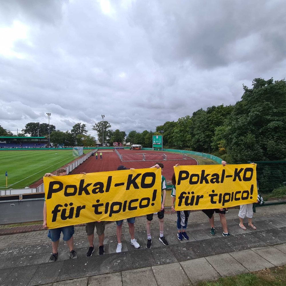 Zwei orangefarbene Banner. Sie haben eine Aufschrift. Die Aufschrift lautet: "Pokal-KO für Tipico". Die Banner sind im Heinz-Dettmer-Stadion in Lohne.