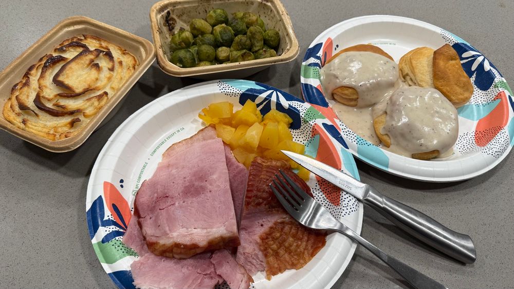 Sliced ham, and pineapple chunks on a paper plate. Small carton of frozen baked mashed potatoes, small carton of frozen baked Brussels sprouts, and a paper plate with 3 premade baked biscuits, and white gravy. 