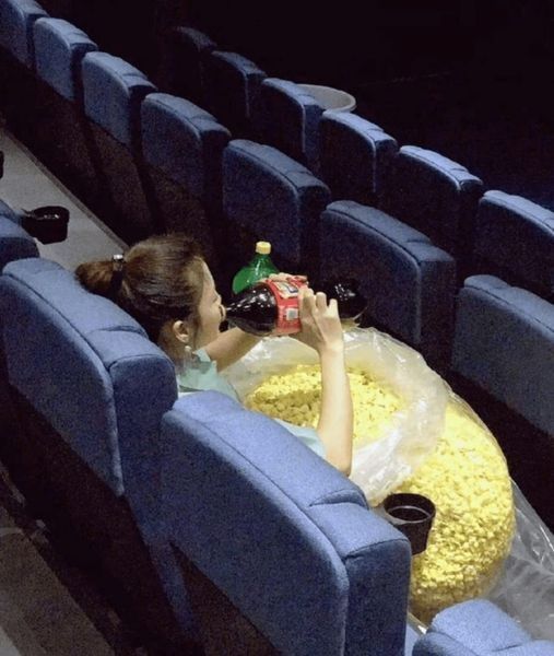 A young girl sitting in an empty movie theater with a bag of popcorn twice the size of her drinking from a 2 liter cola with another one on standby 
