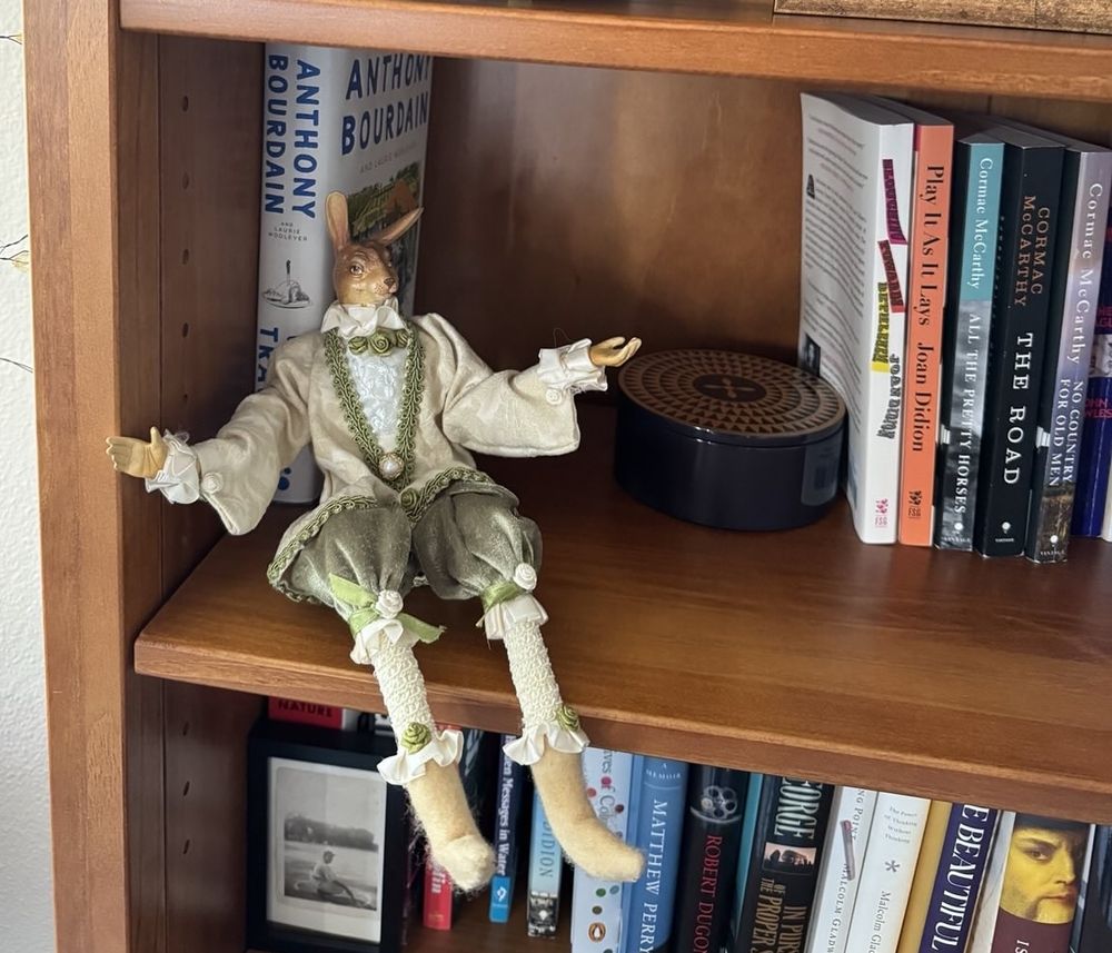 A bunny figurine dressed fancy sitting on a bookshelf. It looks evil. 