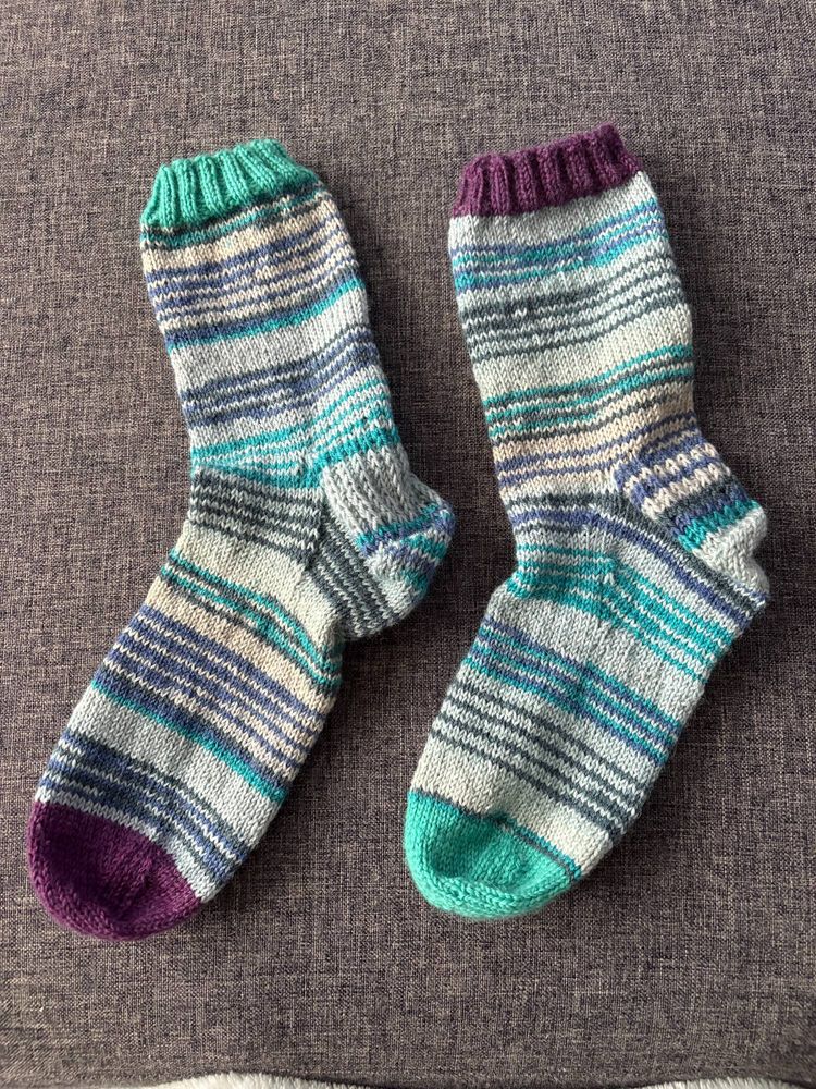 A pair of knit socks made with Lang Yarns SuperSoxx, colourway Artic Ocean. The toe and cuff are different colours, on one socks, the cuff is purple and the toe is turquoise and the other sock is reversed. The toe and cuff are knit with KnitPics Stroll.