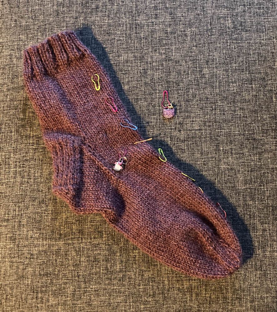 The picture shows my first ever completed sock, in dk weight yarn, in a purple-ish red colour. Some stitch markers are still on as well as a progress keeper with a little cat on it