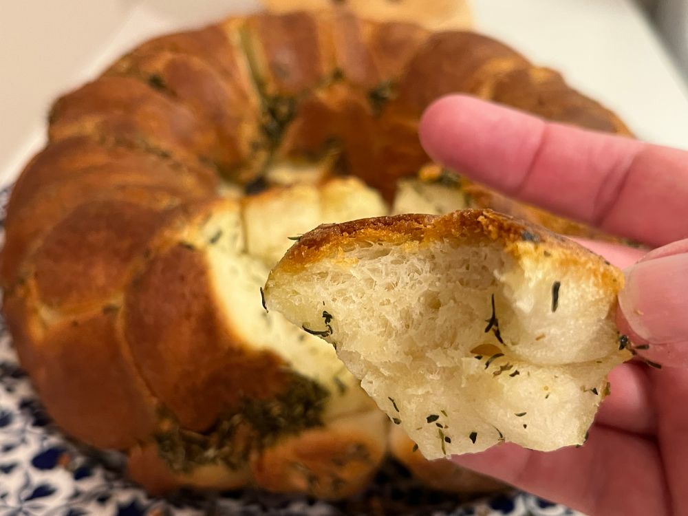 A closeup of a golden brown piece of monkey bread with a bite mark that’s being held in the hand of a white woman. The bread has flecks of green herbs here and there. The rest of the loaf is in the background on a blue and white flower-patterned platter. The bread has a brown crispy outer crust with streaks of more herbs. The crumb looks tender beneath the crust. 