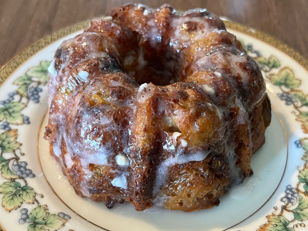 The aforementioned rhubarb cream bundt cake, sitting on a small vintage saucer. It's deep golden brown, covered in a white glaze, with bits of green rhubarb peeking out from the crumb here and there. 