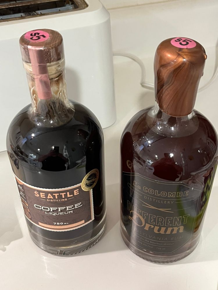 Two hefty sealed and full bottles of dark brown liquor sitting on a white kitchen counter with a white toaster visible in the background. One label reads Seattle Distilling Coffee Liquor and the other label reads La Colombe Distilling Different Drum Pennsylvania Rum. The coffee liqueur is noticeably thicker than the rum, with syrup-like streaks of the concoction running down the inside of the glass. On the top of each sealed lid is a pink price sticker of five dollars. Is it even legal to sell hooch at a yard sale? 