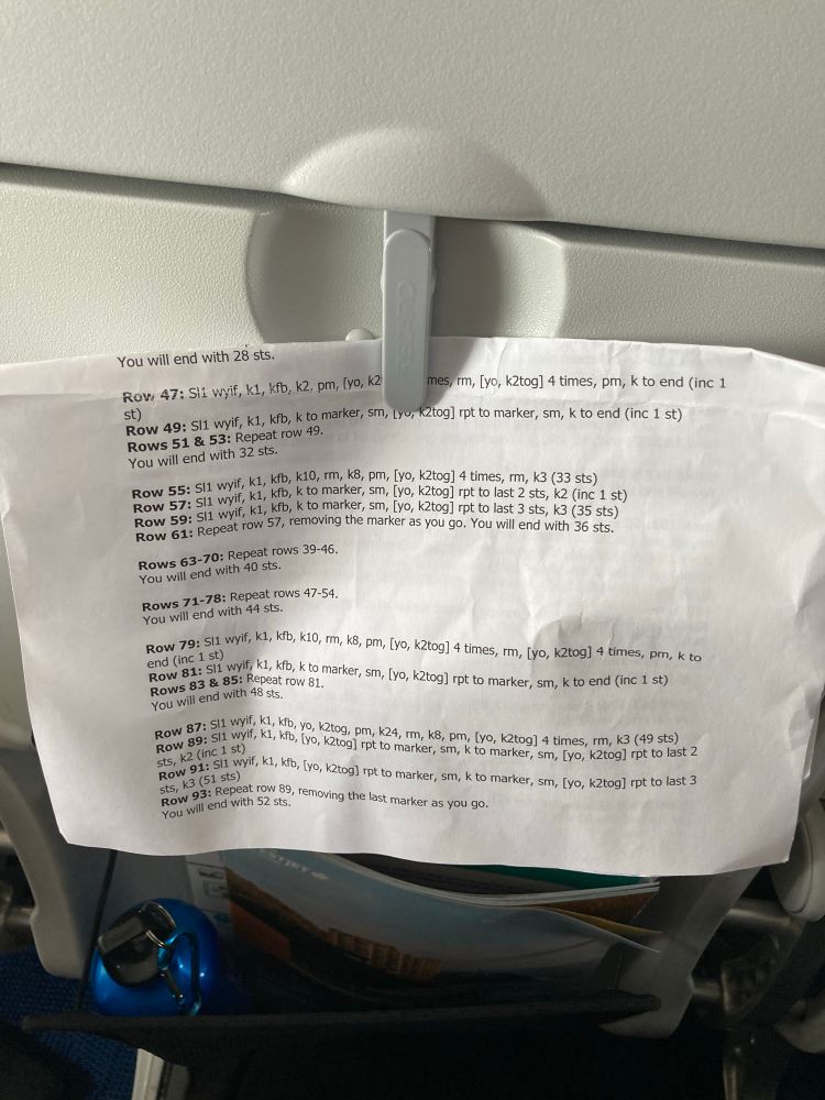 Airline tray table in the upright position, a piece of paper containing a knitting pattern shoved under the latch.