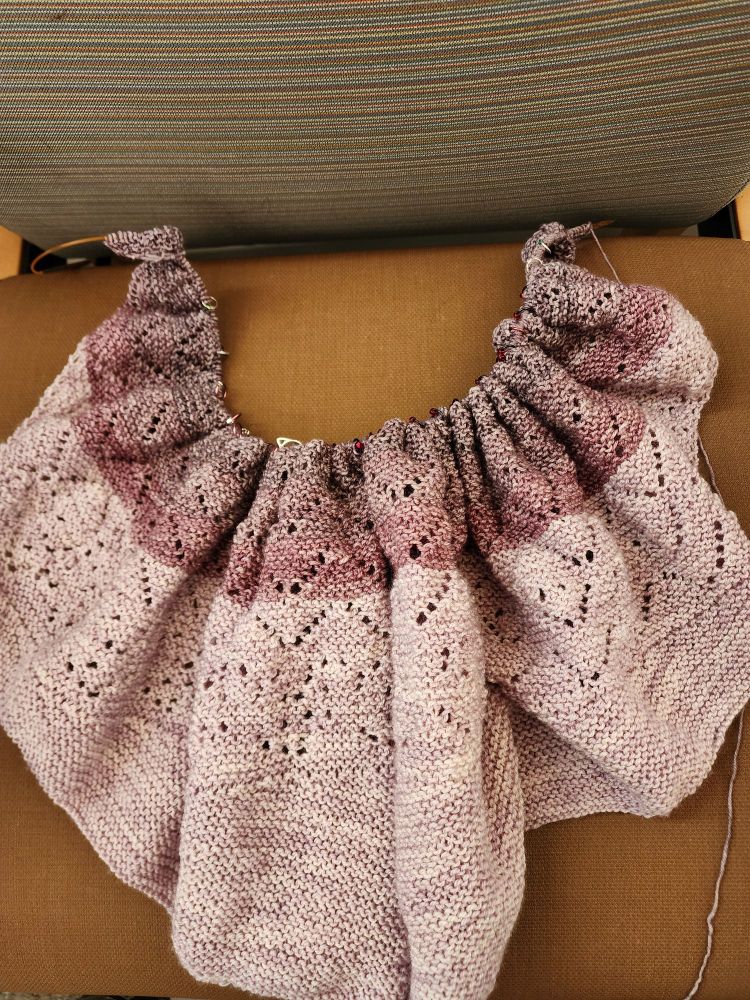 A 3/4 done Allira knitted shawl in pinkish purple colors. The main body is more pink, turning to more purple toward the bottom of the shawl.